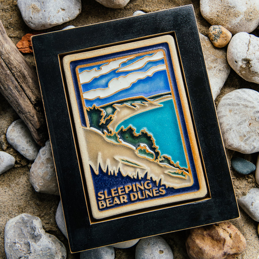 The Sleeping Bear Dunes Tile features the serene Lake Michigan and the sandy beaches and dunes covered on top by bushes and greenery. The words "Sleeping Bear Dunes" line the bottom of the design. The hand painted tile boasts blues and greens which are showcased in a black painted frame.