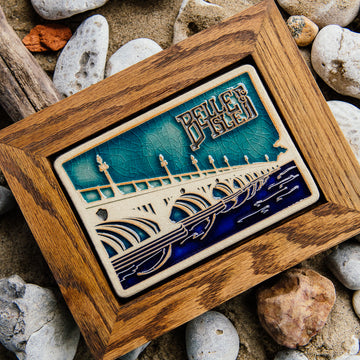 The Belle Isle Bridge Postcard tile features the Macarthur Bridge which connects Belle Isle to mainland Detroit. The words "Belle Isle" float above the bridge in the sky. This tile is hand-painted in a variety of blue glazes which beautifully offset the cool-toned brown of the wood frame.