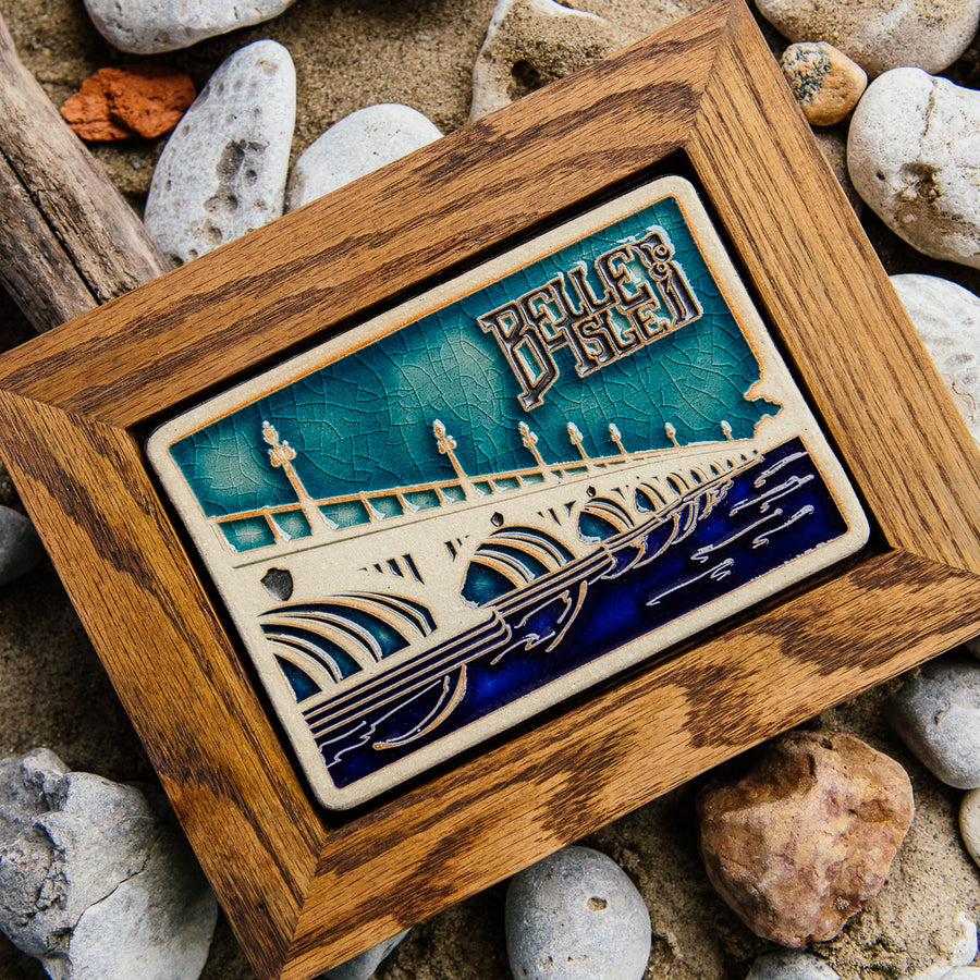 The Belle Isle Bridge Postcard tile features the Macarthur Bridge which connects Belle Isle to mainland Detroit. The words "Belle Isle" float above the bridge in the sky. This tile is hand-painted in a variety of blue glazes which beautifully offset the cool-toned brown of the wood frame.