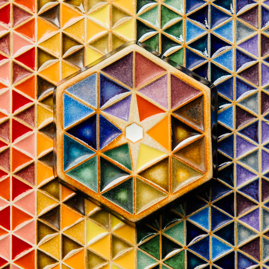 The Hex Paperweight is a thick ceramic tile shaped like a hexagon. The surface of the tile features an interlocking pattern of colorful triangles. In the center of the design is a small white hexagon with a starburst pattern exploding from it. This handpainted paperweight features 24 separate glazes, matte and glossy, in blues, greens, purples, yellows and oranges.