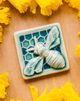 A glossy blue bee tile sits in a bed of daffodils.
