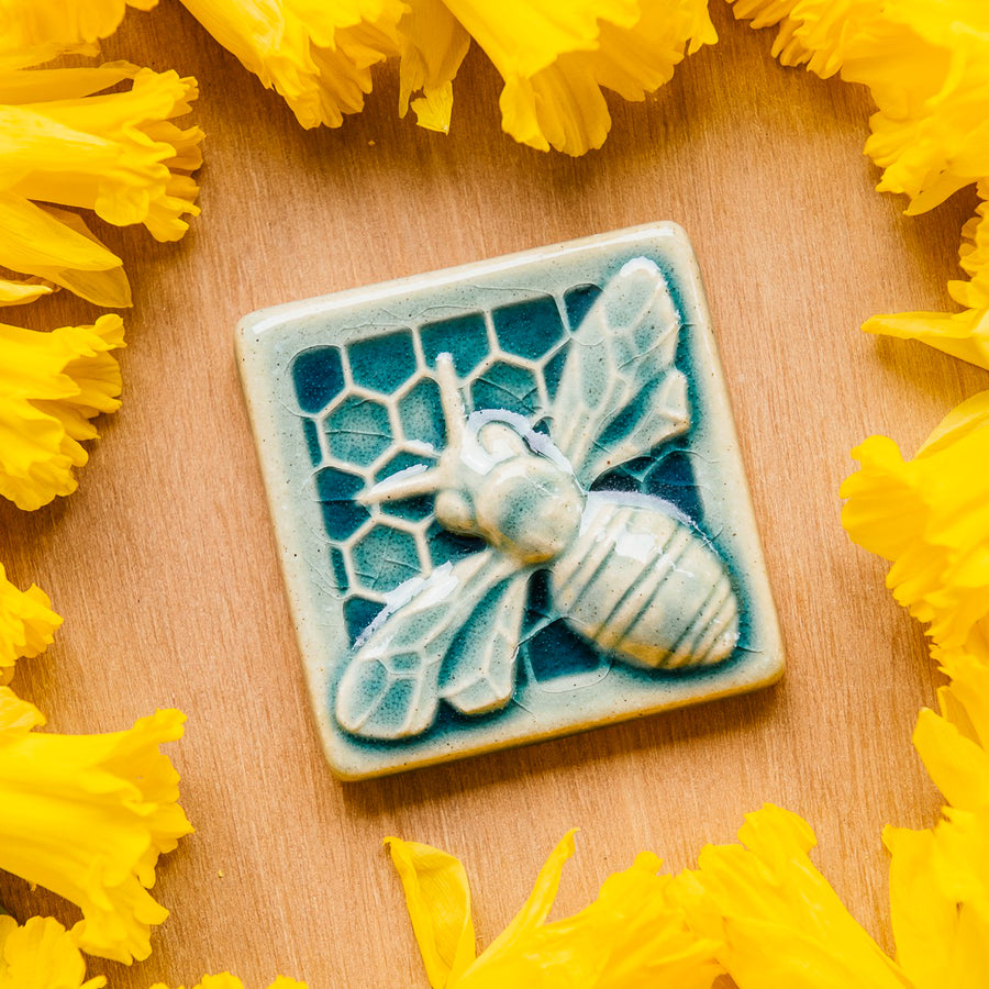 A glossy blue bee tile sits in a bed of daffodils.