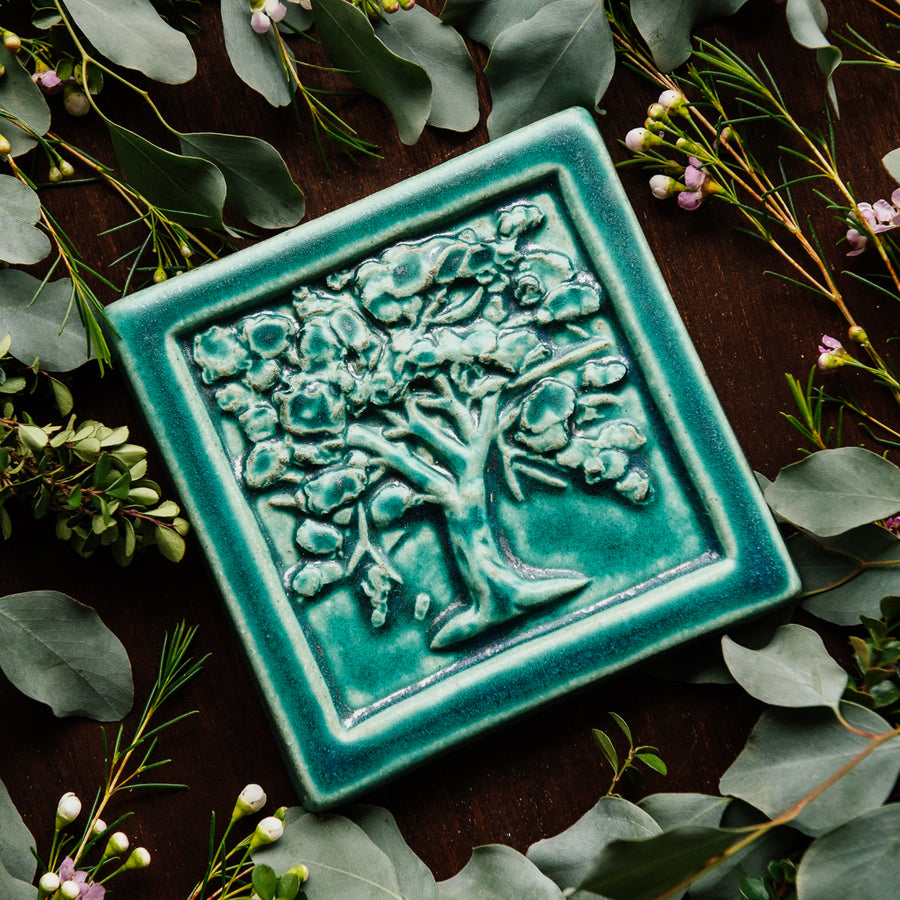 The Oak Tree Tile features a high relief oak tree, covered in layers of abstract leaves. The tile has a thick smooth border. This tile features the matte turquoise Pewabic Blue glaze.