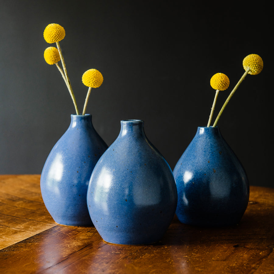 Three Leland Blue Teardrop vases sit together on a wooden table. They carry long yellow flowers with compact round balls on the ends.