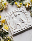 The Lovebirds Tile has a high relief design showing two birds facing one another on a tree branch while pecking at the same berry. There is an ornate border design around it including leaves and twirled lines. This Lovebirds Tile features the creamy white Birch Glaze.