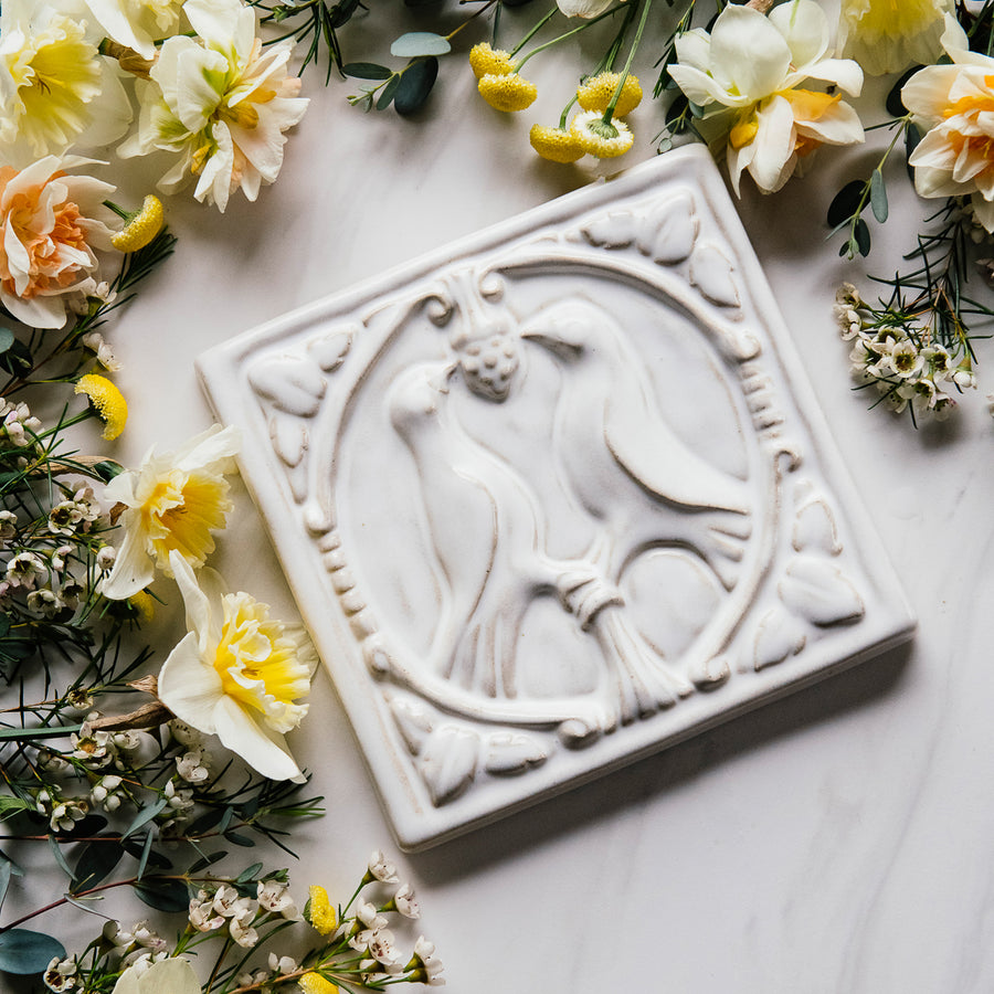 The Lovebirds Tile has a high relief design showing two birds facing one another on a tree branch while pecking at the same berry. There is an ornate border design around it including leaves and twirled lines. This Lovebirds Tile features the creamy white Birch Glaze.