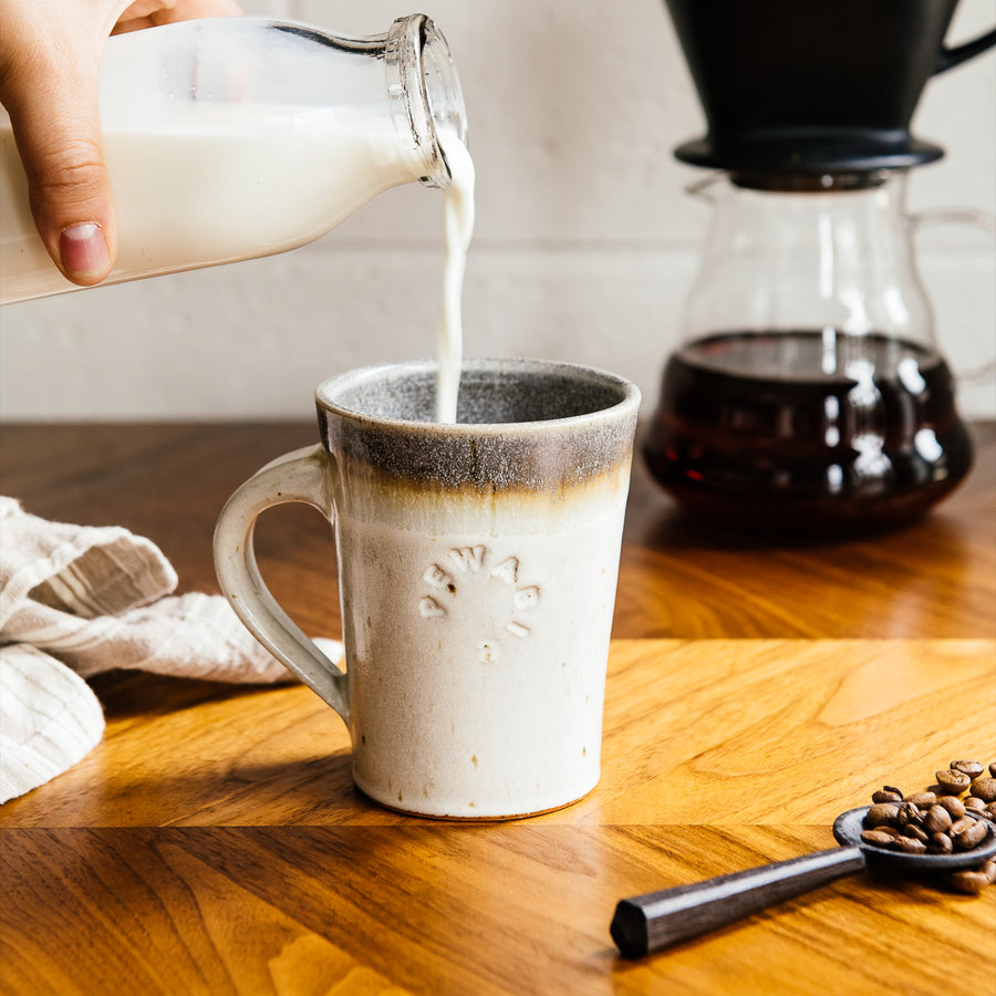 Cafe Mugs have a simple, modern design with smooth, straight sides that slant out. This makes the base slightly smaller than the cup's opening. The round Pewabic logo is stamped onto the side. This cafe mug features the creamy Birch glaze which has natural brown dappling and a grey interior glaze.