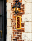 A three digit address frame is attached to a brick house below a porch light. The frame is made of a light blonde wood and holds brick red colored tiles.