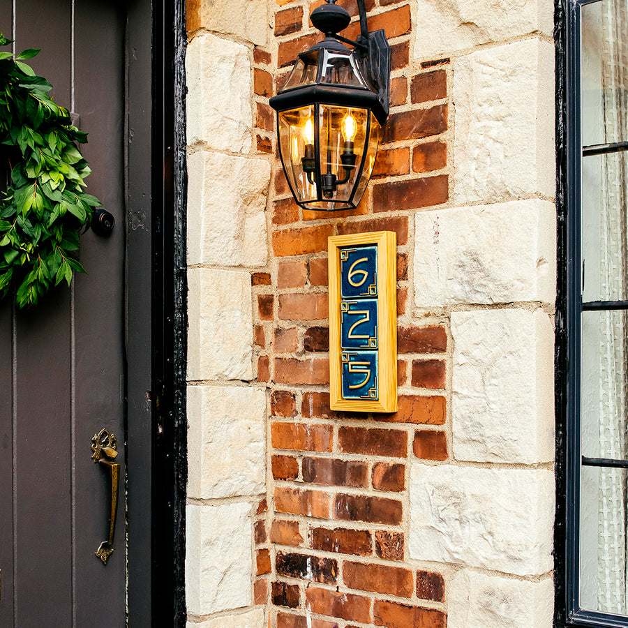 The same three digit address frame is attached to a brick house below a porch light. The frame is made of a light blonde wood and holds peacock blue colored tiles.