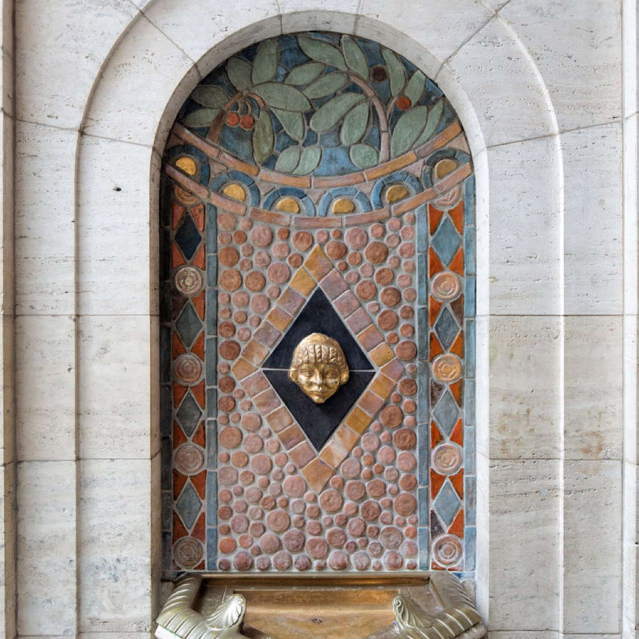 The Pewabic tile drinking fountain sits in a small alcove in a white stone wall. The mosaic tile features circular tiles with harlequin designs on either side. The curved top of the pattern has details of leafy branches with red berries.
