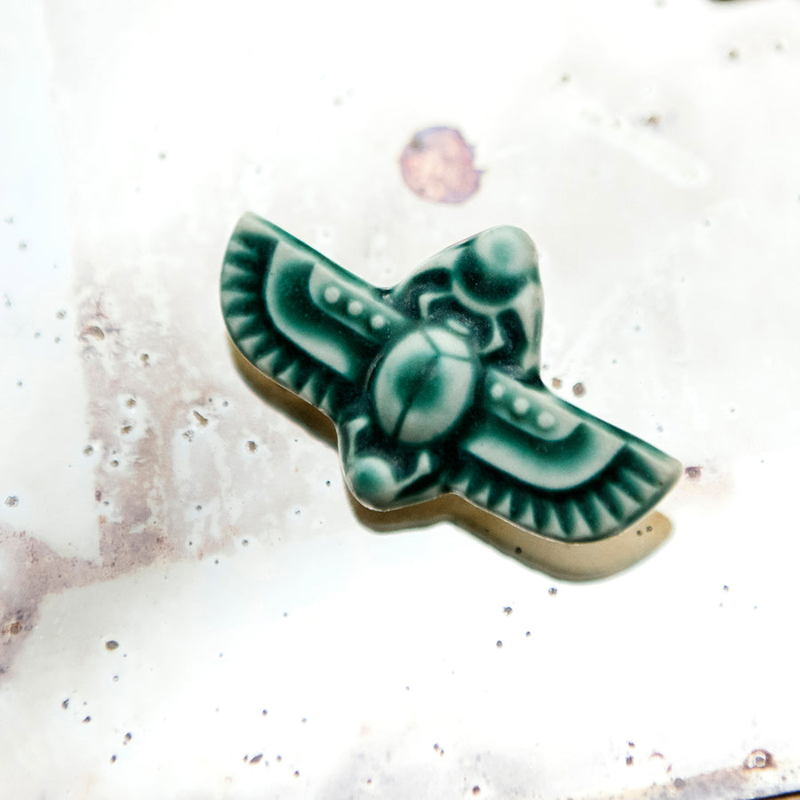 This high relief ceramic pin features a stylized scarab beetle with outstretched wings. This pin has the matte blueish-green Pewabic Green glaze.