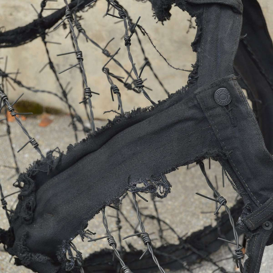 The inner portions of a pair of black jeans have been cut out leaving only the seams to create almost a shell of what once was. In the newly open spaces, barbed wire has been woven across.
