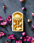 This small rectangular pin has a curved top similar in shape to a stained glass window. Inside there is a delicately carved sunflower facing straight up and a smaller curved bud swirling around its stalk.