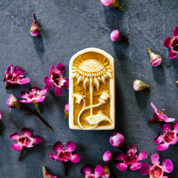 This small rectangular pin has a curved top similar in shape to a stained glass window. Inside there is a delicately carved sunflower facing straight up and a smaller curved bud swirling around its stalk.
