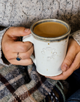 The cafe mug has smooth sides and a simple handle with the word "Pewabic" stamped into the side. The mug is being held by hands wrapped in a cozy sweater.