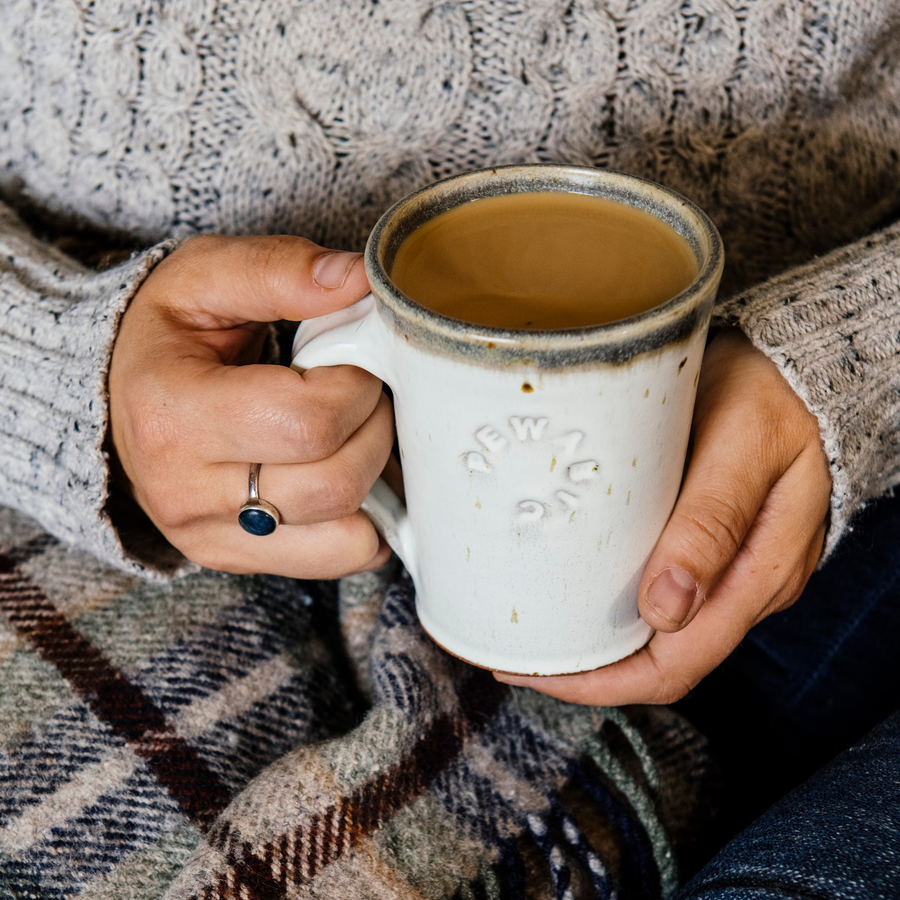 The cafe mug has smooth sides and a simple handle with the word "Pewabic" stamped into the side. The mug is being held by hands wrapped in a cozy sweater.
