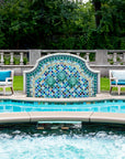 Outdoor poolside fountain in a range of glaze colors that look like the colorful plumes of a peacock. There are three hand-sculpted turtle sculptures at the center of the design in a blue-green glaze. There are two light, wooden lawn chairs on either side of the fountain with blue seat coverings.