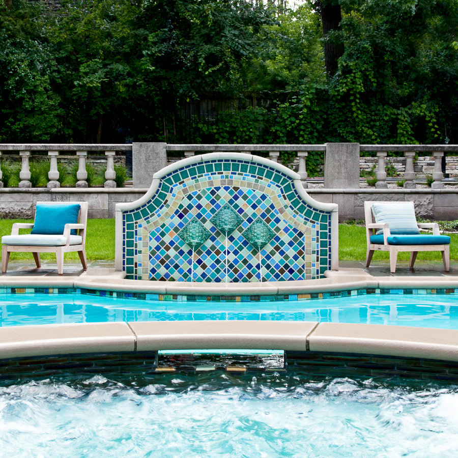 Outdoor poolside fountain in a range of glaze colors that look like the colorful plumes of a peacock. There are three hand-sculpted turtle sculptures at the center of the design in a blue-green glaze. There are two light, wooden lawn chairs on either side of the fountain with blue seat coverings.