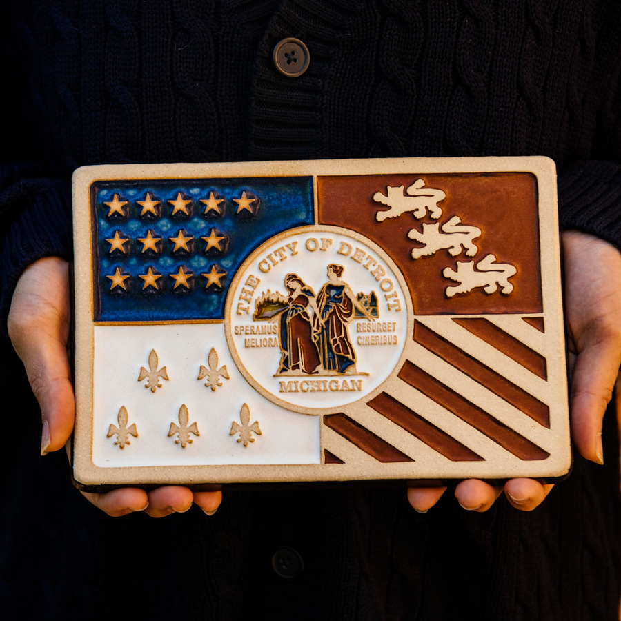 A person in a dark, navy, knit sweater is holding our new hand-painted Detroit Flag Tile.  