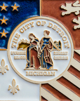 Detail photo of the centerpiece of the Detroit Flag Tile. The circular emblem reads “The City of Detroit” and “Michigan” at the top and bottom. At its center, are two women in red and blue robes. Fire is in the background of the woman on the left and reads “Speramus Meliora” which is latin for “We hope for better things”. The woman on the left is surrounded by a cityscape and reads “Resurget Cineribus” which is “It will rise from the ashes” in latin. 