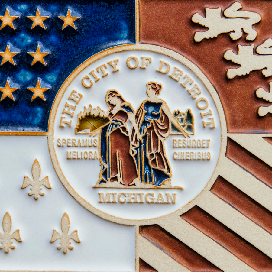 Detail photo of the centerpiece of the Detroit Flag Tile. The circular emblem reads “The City of Detroit” and “Michigan” at the top and bottom. At its center, are two women in red and blue robes. Fire is in the background of the woman on the left and reads “Speramus Meliora” which is latin for “We hope for better things”. The woman on the left is surrounded by a cityscape and reads “Resurget Cineribus” which is “It will rise from the ashes” in latin. 