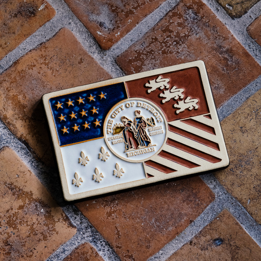 Our hand-painted Detroit Flag Tile is laying on the historic Pewabic tiled floor in our upstairs gallery space. These tiles are a rustic brown and tawny in appearance.