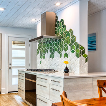 Modern kitchen with range hood against a custom tile backsplash comprised of 4" Rhombus tiles in a gradation of varying green and white glazes. There is a Pewabic Bud Vase with a single yellow flower resting on the glossy white countertop.