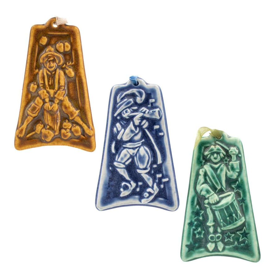 Three colorful embossed ceramic ornaments are on a white background: one shows a man jumping over a stump surrounded by fallen apples, one shows a man dressed in bloomers and a feathered hat as he plays a horn, the last one has a man dressed in Revolutionary War garb as he marches while playing a drum.  