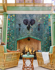 Large scale tile fireplace with high-gloss deep green and blue tiles. The fire is lit and there is a metal elephant head at the center of the design alongside two metal torches. There are light green pillars on each side of the fireplace. There is a wicker furniture set with sea-foam green cushions just within frame. There is a woman enjoying a glass of wine in the chair to the left of the fireplace.