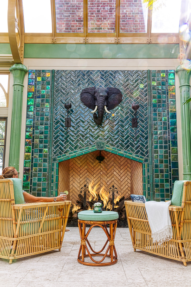 Large scale tile fireplace with high-gloss deep green and blue tiles. The fire is lit and there is a metal elephant head at the center of the design alongside two metal torches. There are light green pillars on each side of the fireplace. There is a wicker furniture set with sea-foam green cushions just within frame. There is a woman enjoying a glass of wine in the chair to the left of the fireplace.