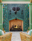 Large scale tile fireplace with high-gloss deep green and blue tiles. The fire is lit and there is a metal elephant head at the center of the design alongside two metal torches. There are light green pillars on each side of the fireplace. There is a wicker furniture set with sea-foam green cushions just within frame.  