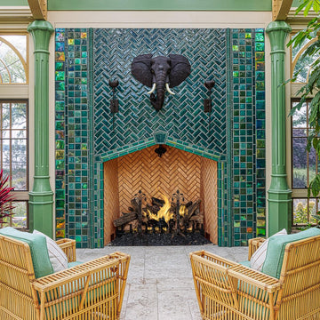 Large scale tile fireplace with high-gloss deep green and blue tiles. The fire is lit and there is a metal elephant head at the center of the design alongside two metal torches. There are light green pillars on each side of the fireplace. There is a wicker furniture set with sea-foam green cushions just within frame.  