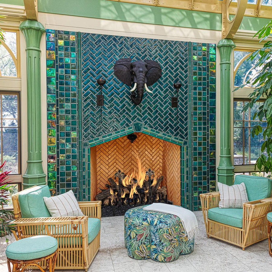 Large scale tile fireplace with high-gloss deep green and blue tiles. The fire is lit and there is a metal elephant head at the center of the design alongside two metal torches. There are light green pillars on each side of the fireplace. There is a wicker furniture set with sea-foam green cushions just within frame.  