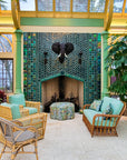 Large scale tile fireplace with high-gloss deep green and blue tiles. The fire is lit and there is a metal elephant head at the center of the design alongside two metal torches. There are light green pillars on each side of the fireplace. There is a wicker furniture set with sea-foam green cushions just within frame. Tropical plants are also coming into the frame of the photo.