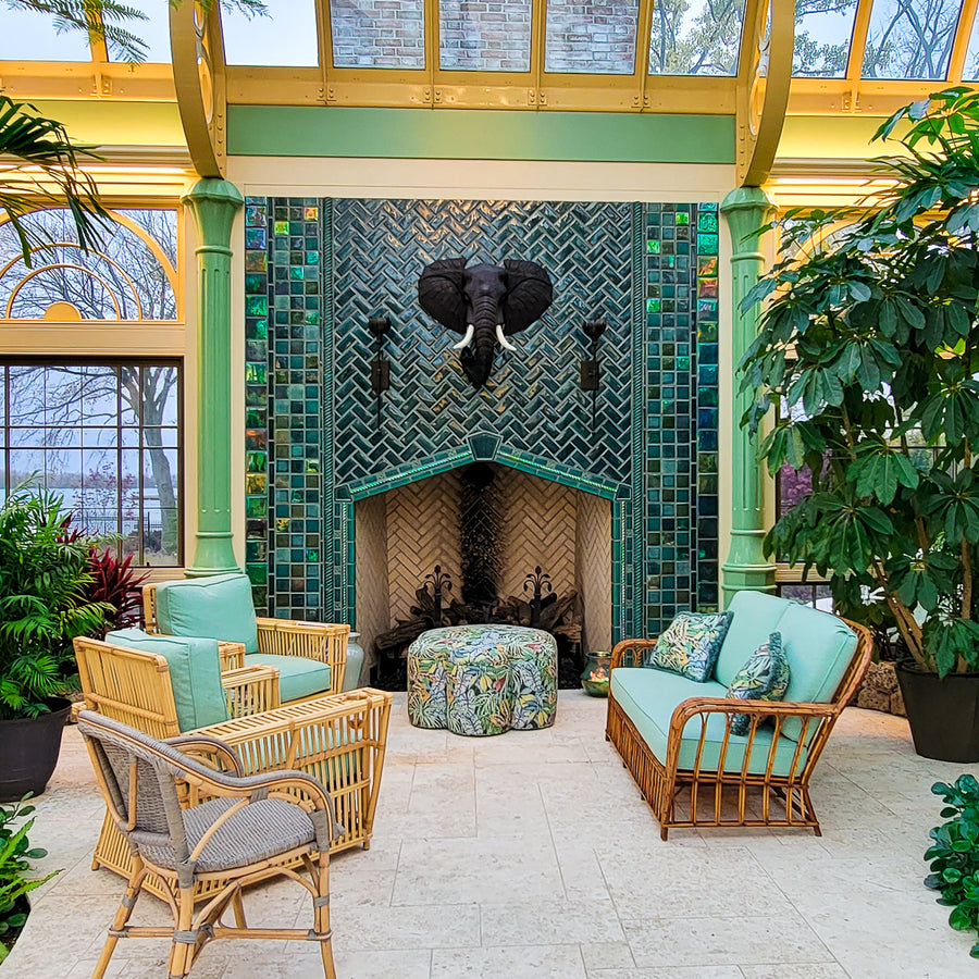 Large scale tile fireplace with high-gloss deep green and blue tiles. The fire is lit and there is a metal elephant head at the center of the design alongside two metal torches. There are light green pillars on each side of the fireplace. There is a wicker furniture set with sea-foam green cushions just within frame. Tropical plants are also coming into the frame of the photo.