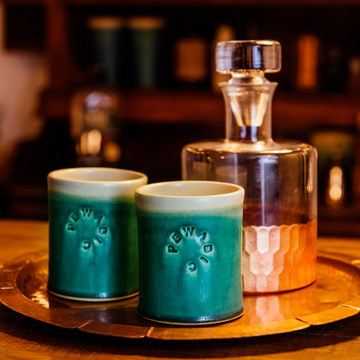The Rocks Cups are short, cylindrical cups with smooth, straight sides. The Pewabic logo is stamped onto the side. There are two glazes featured on each cup - one is on the outside, covering the logo while the other glaze is on the inside of the cup and spills over creating a half inch ring around the outer lip of the cup. 