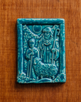 The Shepherds Tile features a man dressed in robes with his hand on the shoulder of a young boy. A sheep stands at their feet and the moon looks down at them through a large hole in the brick wall behind them. This tile features the matte turquoise Pewabic Blue glaze.
