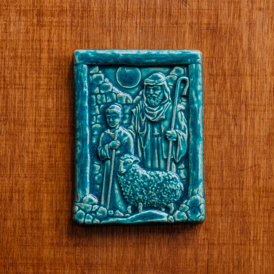 The Shepherds Tile features a man dressed in robes with his hand on the shoulder of a young boy. A sheep stands at their feet and the moon looks down at them through a large hole in the brick wall behind them. This tile features the matte turquoise Pewabic Blue glaze.