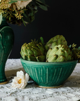 A Pewabic Green classic bowl sits on a lace table cloth. Large, scaly artichokes overflow from it.