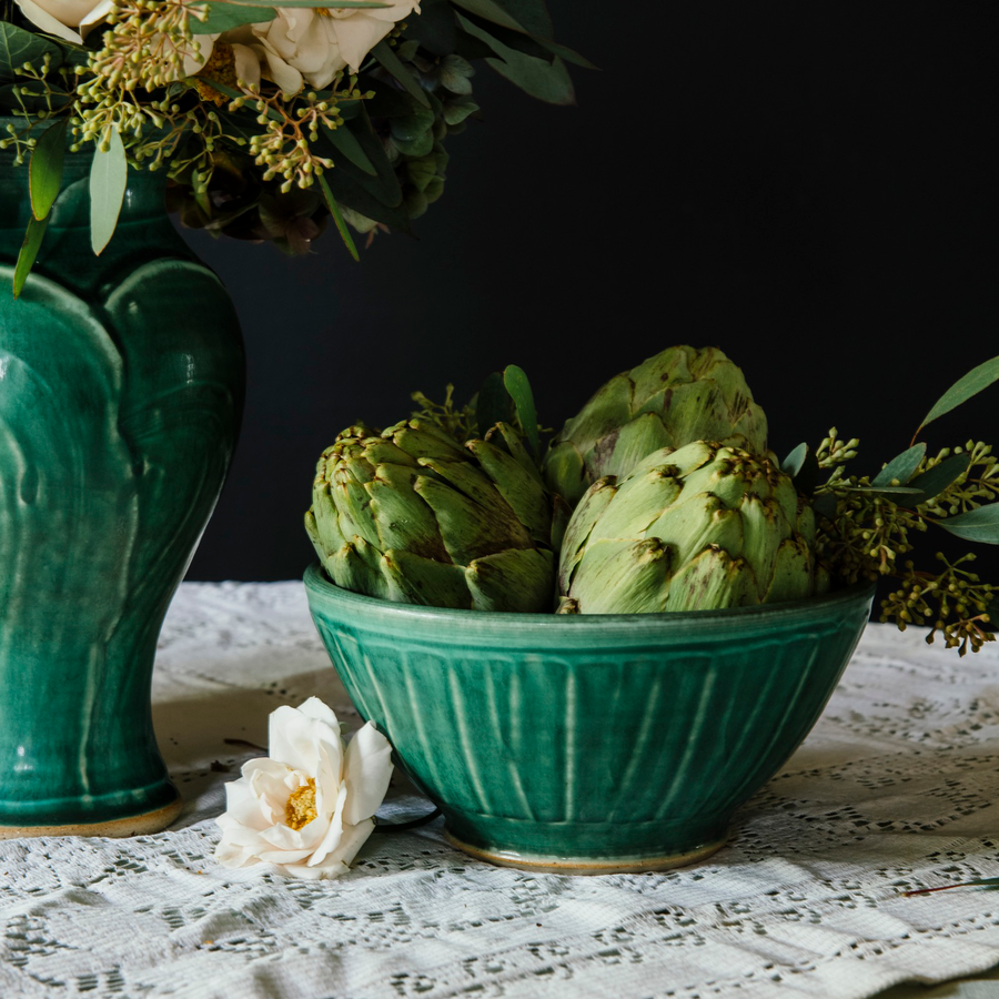A Pewabic Green classic bowl sits on a lace table cloth. Large, scaly artichokes overflow from it.