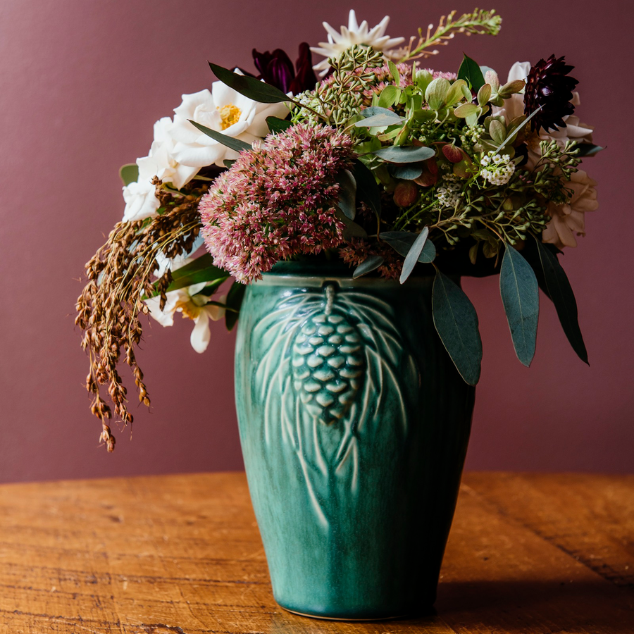 This Pinecone vase features the matte blueish-green Pewabic Green glaze, filled with a fall inspired flower arrangement featuring burgundy, rust, peach, and creme flowers.