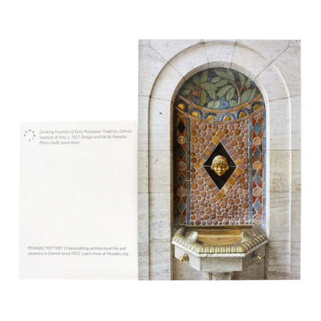 The DIA Fountain Postcard features an image of a historic drinking fountain. The fountain and handles are a brushed metallic gold and above it is Pewabic tile mural tucked into an alcove of a marble wall. The intricate mural design has circular tiles leading up to branches with red berries at the top. In the center of the installation is a golden face of a man.