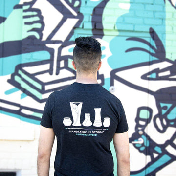 The back of the Handmade in Detroit Tee features a drawing of a line of Pewabic vases - two petites, a Calla Lily vase, a Snowdrop Vase and a Step Vase. They are all in white with the words "Handmade in Detroit Pewabic Pottery below them."