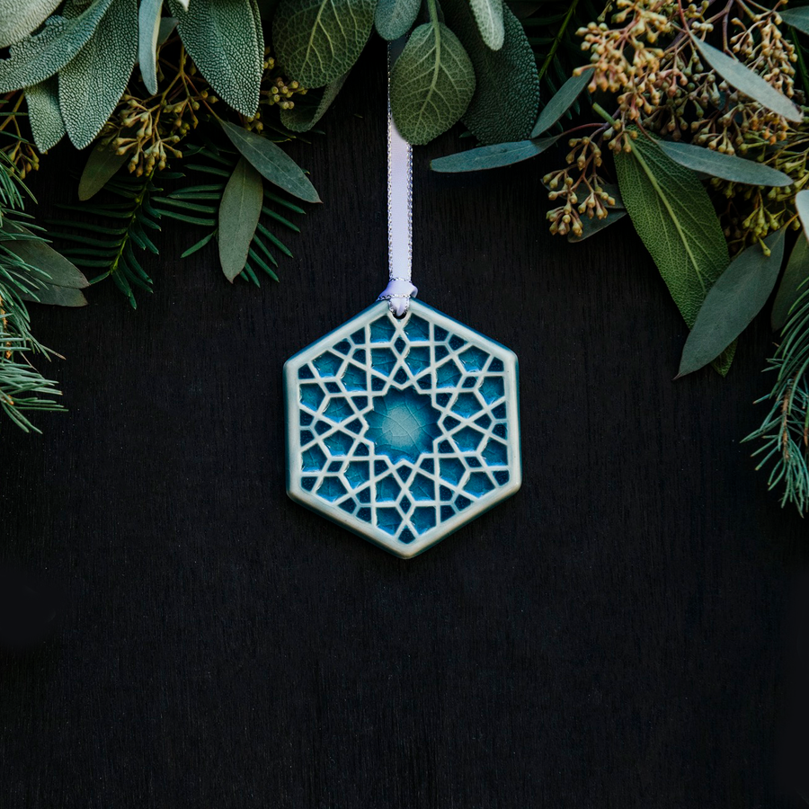 Our 2022 Snowflake Ornament named “Ice” in a high-gloss, light-blue, Glacier Gloss glaze against a black backdrop. The ornament hangs from a white ribbon lined with silver against a surround of wintery greenery.