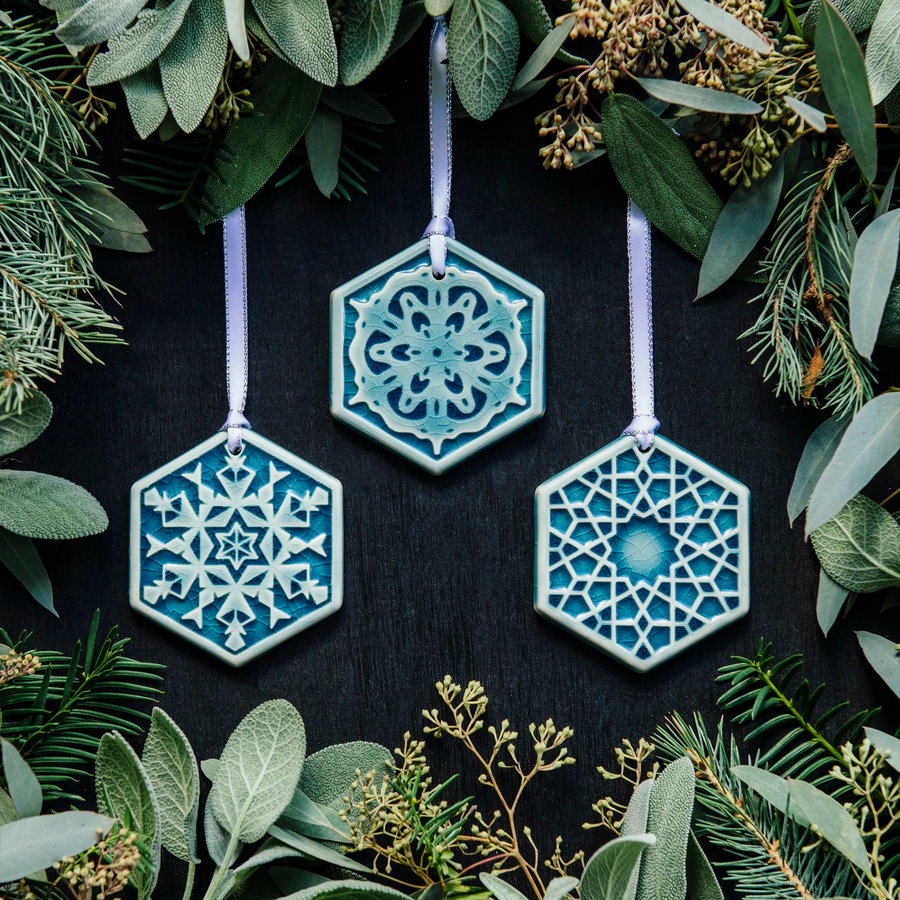 Photo of all three Snowflake Ornament designs chosen for our 2022 Winter Collection. From left to right: “Frosty”, “Snowfall”, and “Ice”. All ornaments are surrounded by winter greenery against a black background. 