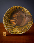 Style A is an irregularly shaped rounded plate. It has organic ridges around its lip mimicking a spiral shell. The glaze starts in a rusty brown in the center and becomes more yellowy near its edges.