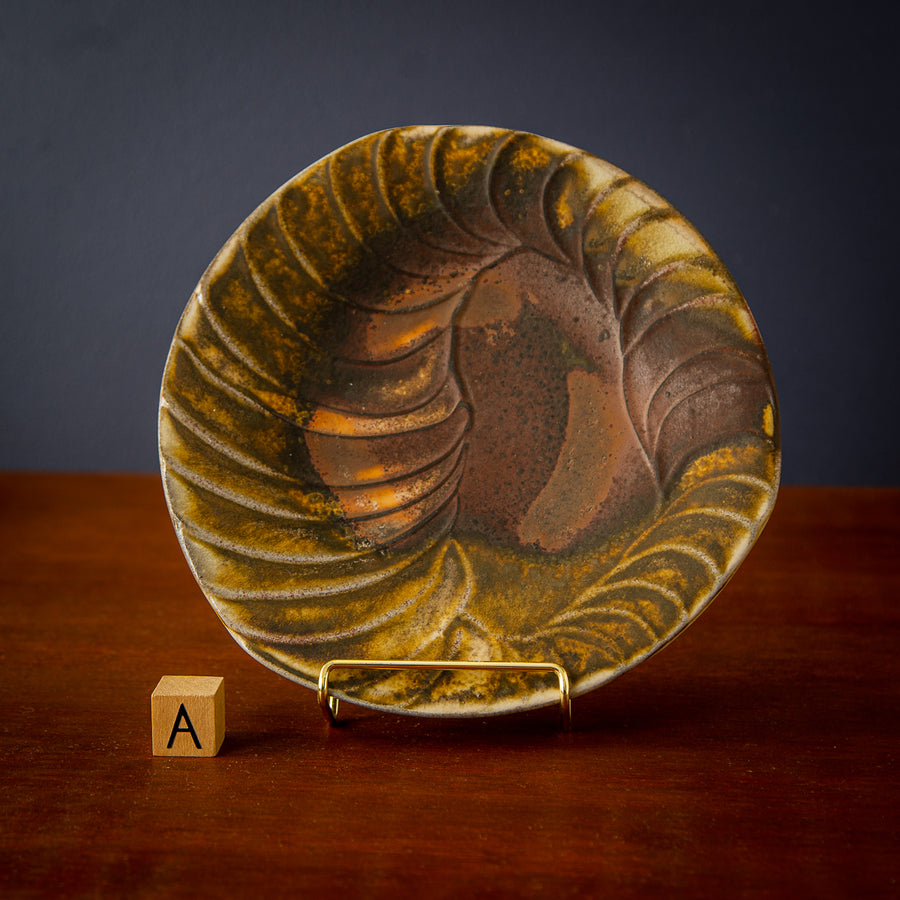 Style A is an irregularly shaped rounded plate. It has organic ridges around its lip mimicking a spiral shell. The glaze starts in a rusty brown in the center and becomes more yellowy near its edges.