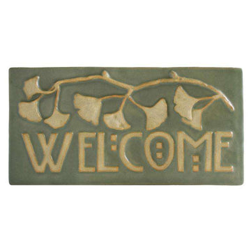 The Ginkgo Welcome Tile features the word "Welcome" in Arts and Crafts style font with a branch of ginkgo leaves above it. The leaves and wording are scraped giving them a creamy off-white color with a pale green glazed background.