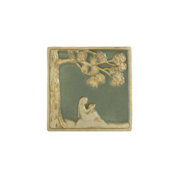 The Girl Reading Tile features a young girl with long hair sitting in profile under a large tree. A branch is above her, giving protection while she reads from a book. The tree and girl are scraped giving them a creamy off-white color while the background is a pale green glaze.