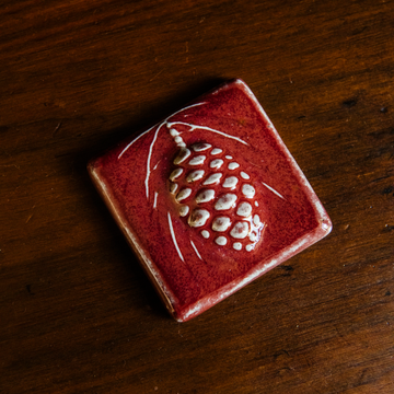 The ceramic Pinecone Tile has a high relief design with one large pinecone in the center and long spindly pine needles framing it.